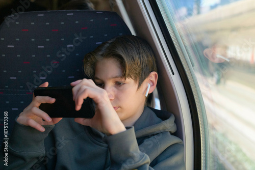 A teenager wearing wireless earbuds focuses intently on his smartphone while sitting by the window on a train. photo