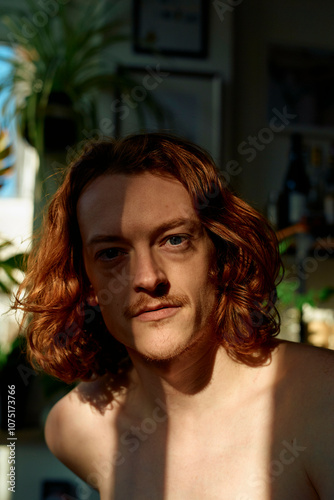 Half-lit portrait of a shirtless man with shoulder-length wavy hair in a room with plants photo