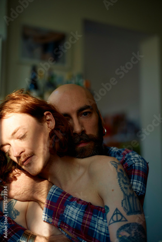 Bald man with beard and shirt embraces a shirtless young man with red hair as sunlight filters through a window, casting shadows on them. photo