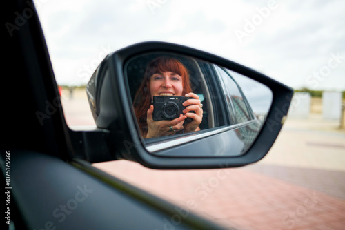 A smiling woman takes a photo of herself in the side mirror of a car. photo