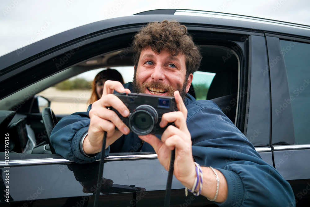 Smiling man with curly hair taking a photo from a car window with a digital camera Smiling man with curly hair taking a photo from a car window with a digital camera