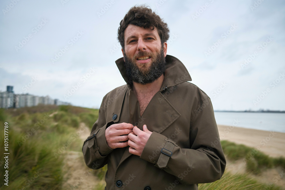 Smiling bearded man buttoning up his coat on a cloudy beach day with city skyline in the background, Belgium Smiling bearded man buttoning up his coat on a cloudy beach day with city skyline in the background, Belgium