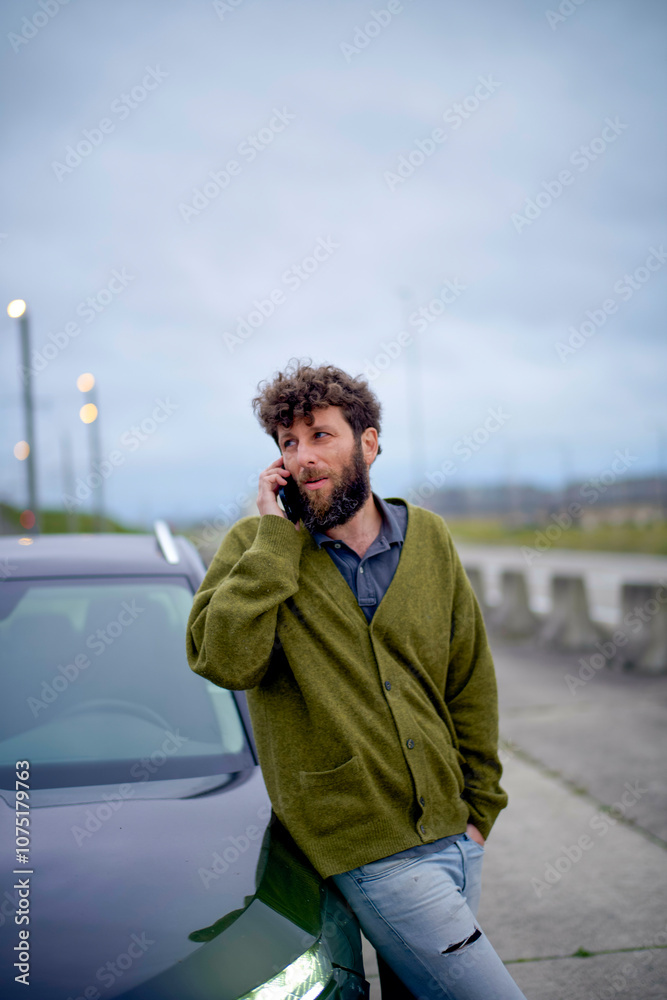 Man with curly hair talking on his phone while leaning against a car outdoors at twilight. Man with curly hair talking on his phone while leaning against a car outdoors at twilight.