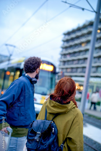 Two individuals wait at a tram station during dusk, with one carrying a blue backpack and the other wearing headphones, Belgium photo