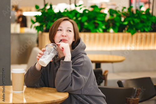 Middleaged woman sitting at caffee in shopping mall and drinking milkshake, while using mobile phone or looking away