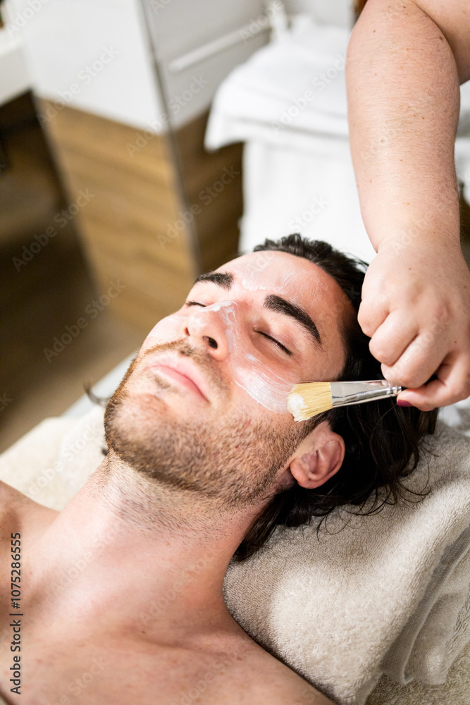 Obraz premium Vertical photo of a handsome bearded man in his 30s and 40s is inside a spa.The male is lying on a table while undergoing a facial massage with a brush.Massages with brushes and oil.