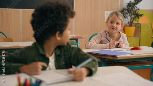 Multiracial diverse little classmates multiethnic students pupils two children kids Caucasian girl help African American boy suggest correct answer to exam writing test talking in school class lesson