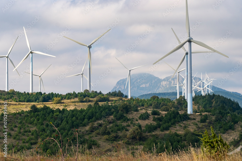A collection of wind turbines gracefully stand atop rolling hills, with majestic mountains forming a serene backdrop under a slightly cloudy sky, illustrating green energy in Navarra Spain A collection of wind turbines gracefully stand atop rolling hills, with majestic mountains forming a serene backdrop under a slightly cloudy sky, illustrating green energy in Navarra Spain