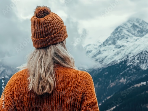 a woman clad in a cozy orange sweater and a beanie gazes thoughtfully at distant mountains, her silhouette framed against a stunning backdrop of snow-capped peaks