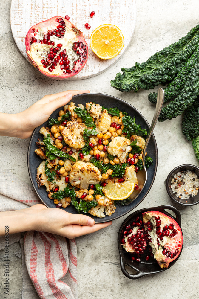 Cooking Roasted Cauliflower and roasted chickpeas salad, with pomegranate, kale, with spices, oil and lemon dressing. Vegetable winter season food on a light background. Vertical image.