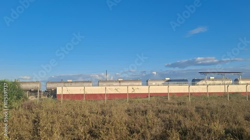 Freight train moving past an industrial building with a tall grass foreground, framed against a clear blue sky on a sunny day