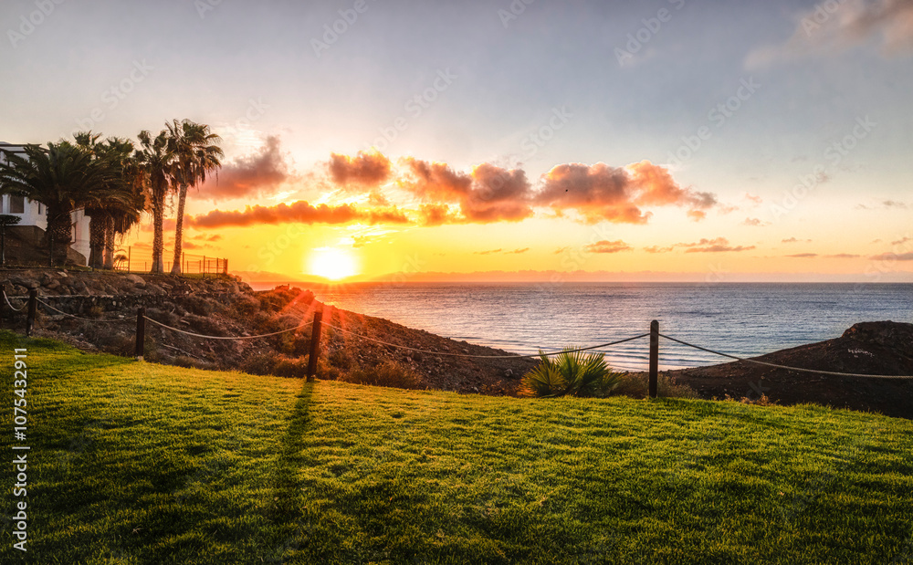 Eine Sonnenaufgang in einer Hotelanlage auf Fuerteventura. Palmen und sanfte Hügel vor dem Meerblick. Saftiges grünes Gras im Vordergrund. Eine Sonnenaufgang in einer Hotelanlage auf Fuerteventura. Palmen und sanfte Hügel vor dem Meerblick. Saftiges grünes Gras im Vordergrund.
