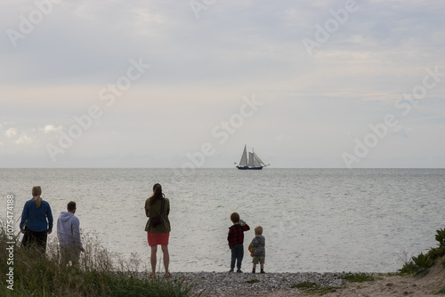 Backview of a family with two small kids standing at the beach watching a sailing vessel passing by photo