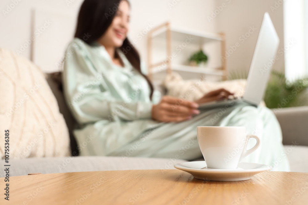 Cup of coffee on table against woman in pajamas working with laptop at home, closeup Cup of coffee on table against woman in pajamas working with laptop at home, closeup