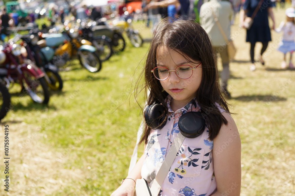 Fototapeta premium Little girl in glasses and headphones having fun against the background of old cars and motorcycles. Concept of children's travel, activity, fun and getting new impressions