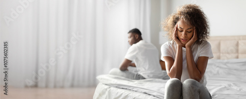 A black woman sits on the edge of a bed, holding her head in distress, while a man sits apart from her, symbolizing emotional distance and communication issues in their relationship.