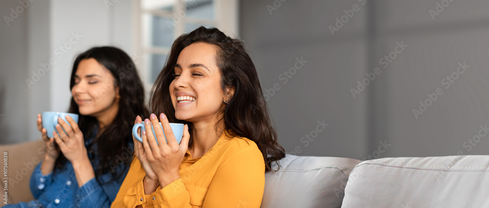 Two women are sitting comfortably on a sofa in a cozy living room, savoring warm drinks. They appear relaxed and content, enjoying each other's company and the peaceful atmosphere. Two women are sitting comfortably on a sofa in a cozy living room, savoring warm drinks. They appear relaxed and content, enjoying each other's company and the peaceful atmosphere.