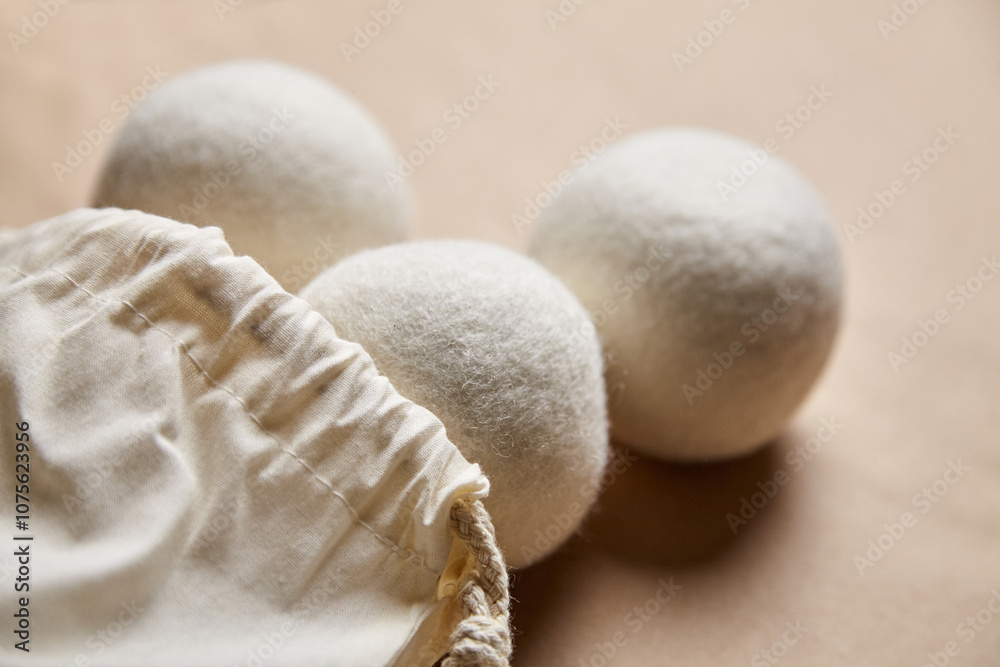 Close-up of three white wool dryer balls next to an open cotton drawstring bag on a beige background, ideal for sustainable laundry routines