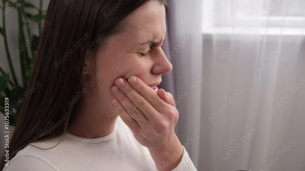 Close up of young woman feel terrible toothache after drink cold water. Female sitting on sofa at home touching cheek, feel hurt and suffering from sensitive tooth ache. Pain and cavities concept Close up of young woman feel terrible toothache after drink cold water. Female sitting on sofa at home touching cheek, feel hurt and suffering from sensitive tooth ache. Pain and cavities concept