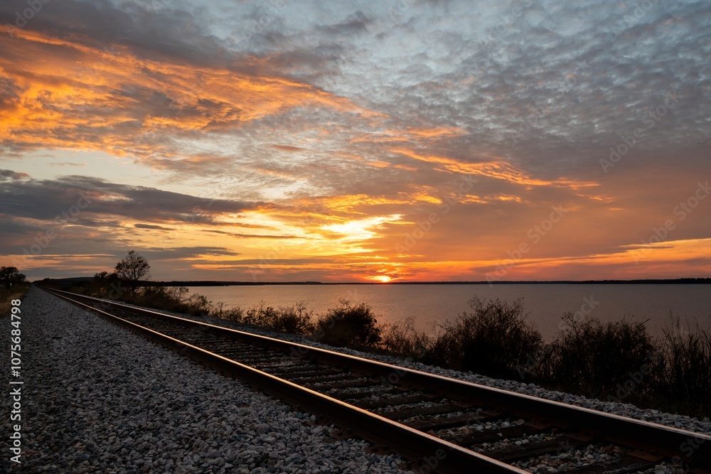 Railroad tracks leading off into the distance along the shore of a lake as the sunset fills the sky with fiery yellow and orange clouds. Railroad tracks leading off into the distance along the shore of a lake as the sunset fills the sky with fiery yellow and orange clouds.