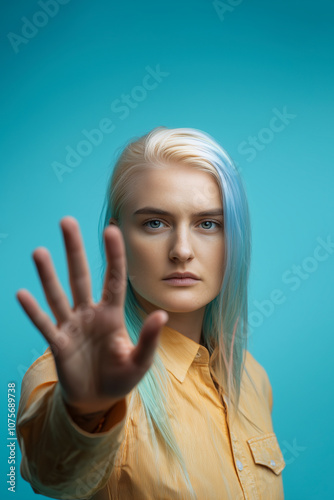 Young transgender woman with platinum blonde and pastel blue hair, raising a hand in a stop gesture to protest transgender discrimination. Mustard yellow shirt. Teal background. Aware poster. photo