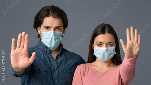 Social Distancing Concept. Portrait of serious millennial couple in protective medical masks showing stop gesture with open palm over light background