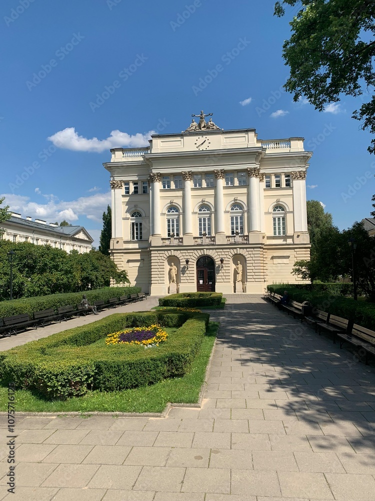 Fototapeta premium Front view of an elegant historical building with classic architecture surrounded by a manicured garden under a clear blue sky. Ideal for themes of history, architecture, and travel.