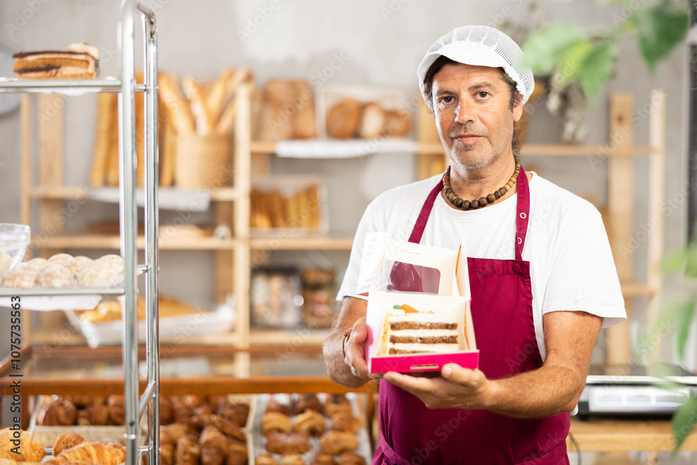 Positive middle-aged salesman offering a piece of cake packed in box in bakery Positive middle-aged salesman offering a piece of cake packed in box in bakery