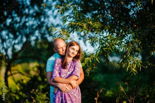 A happy mid-adult couple is tenderly hugging outdoors in a green park.The man and woman are smiling cheerfully while looking directly at the camera.