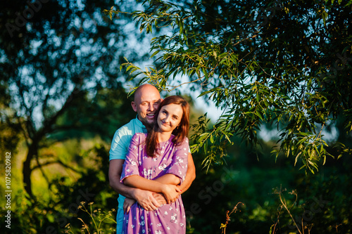 A happy mid-adult couple is tenderly hugging outdoors in a green park.The man and woman are smiling cheerfully while looking directly at the camera.