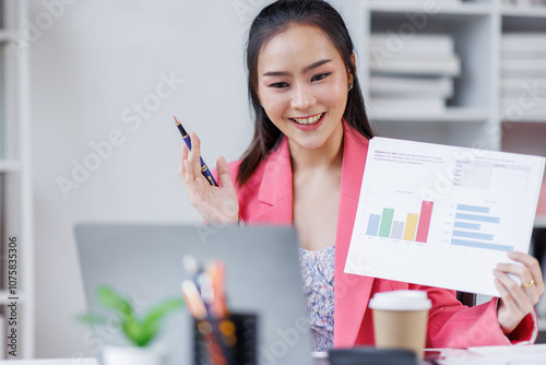 Asian business woman shows documents with clipboard graphs holds in her hand, looks at the camera loptop, smiles. Sitting at a desk with a laptop in a modern office.
 photo