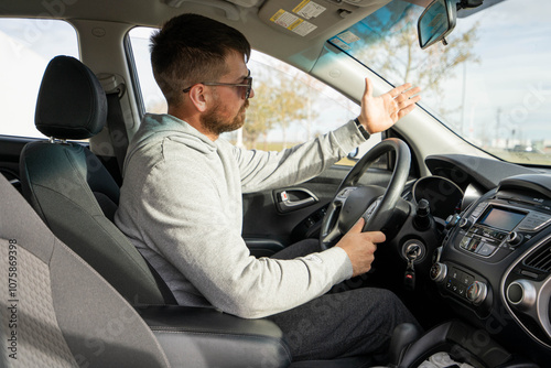 Young Caucasian man with beard point his hand towards windshield as he stays long time in the traffic and loosing his patience already