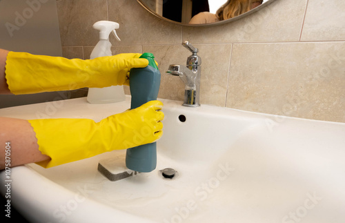 Woman in yellow gloves opening green bottle with household cleaning liquid to clean a sink in the bathroom. Cleaning service in process.