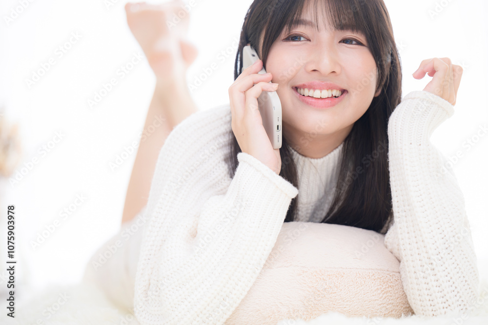 うつ伏せでスマホを通話するニットを着た若いかわいい笑顔の女子のクローズアップ　明るい白いかわいいコミュニケーションなどのイメージ