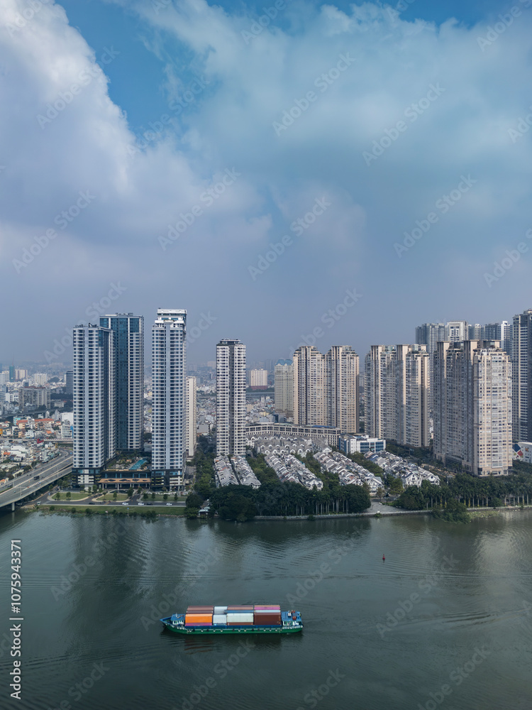 Fototapeta premium Freighter with containers on river against background of modern high rise and villas in new residential area with dramatic sky.