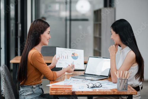 Two professional women discussing financial charts in a contemporary office environment, showcasing teamwork and business strategy.