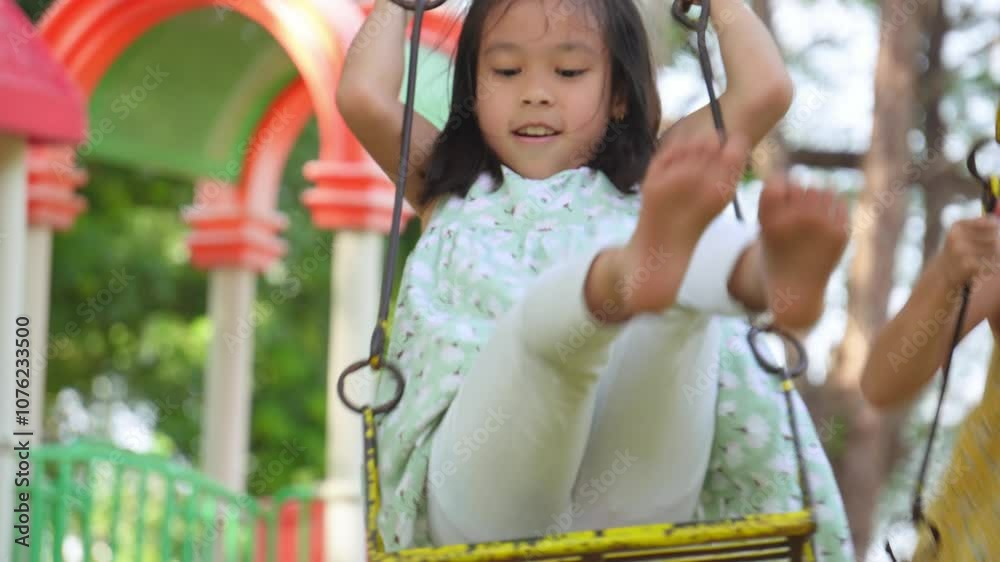 Cute sisters are having fun playing on the swings in a playground in a park. Children playing in the playground during summer vacation. Cute sisters are having fun playing on the swings in a playground in a park. Children playing in the playground during summer vacation.