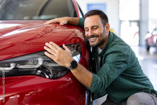 Happy young man hugging his new car in showroom. Satisfied guy with closed eyes embracing the hood of the automobile. Dreaming man lying on car bonnet hugging it. photo