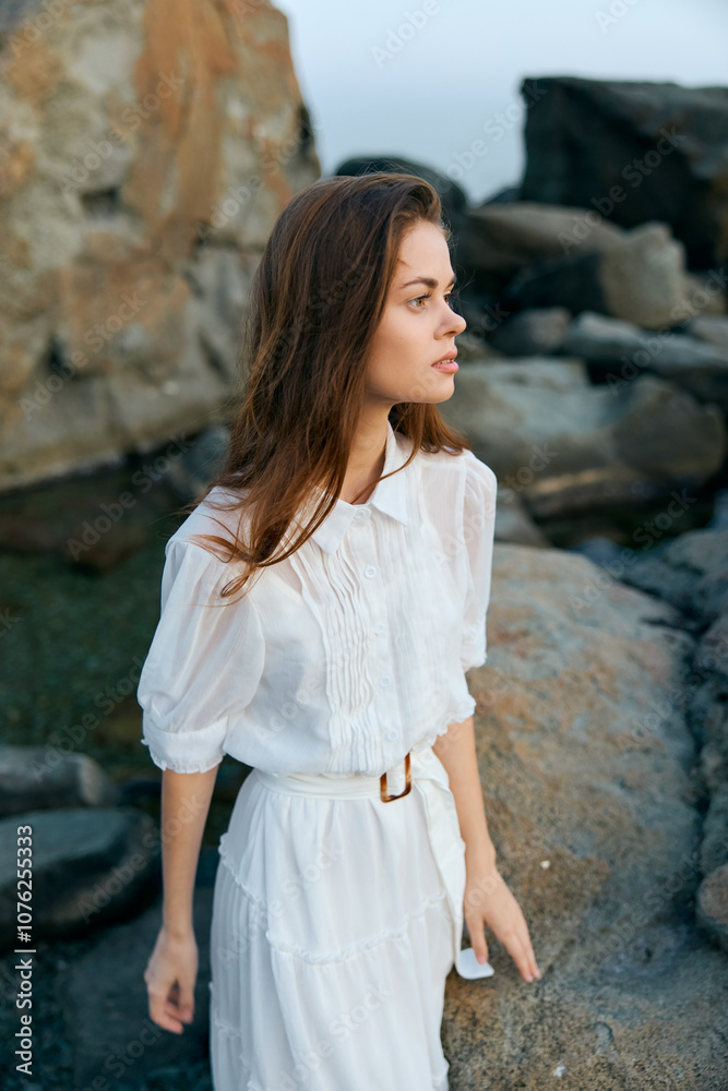 Majestic woman in flowing white dress standing on rocky shoreline against backdrop of ocean waves