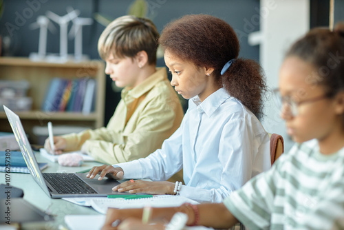 Side view of diverse group of children sitting in row in school classroom with focus on young African American girl using computer copy space