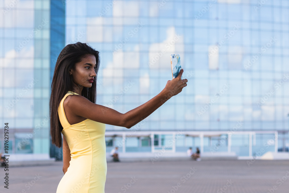 Fototapeta premium Indian woman in yellow dress taking a selfie with smartphone in front of a modern glass building