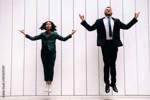 Smiling businessman jumping and doing yoga with businesswoman in front of white wall photo