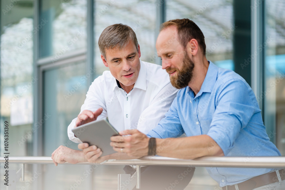 Colleagues leaning on railing in office building discussing business strategy using digital tablet Colleagues leaning on railing in office building discussing business strategy using digital tablet