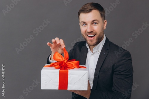 Bearded happy smiling man in business suit holding a gift and unwrapping it on a gray background, valentines day, christmas and new year concept