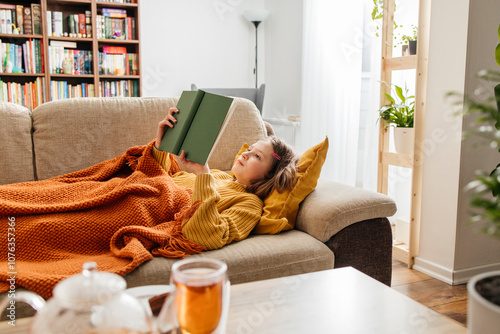 Girl reading book and lying with knitted blanket on sofa at home