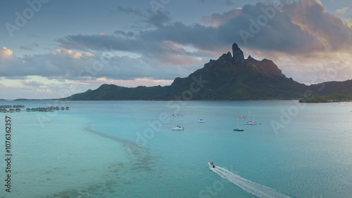 Speedboat sailing turquoise water near tropical island, green mountains and bungalows on water at sunset. Bora Bora, French Polynesia exotic paradise. Perfect summer travel vacation aerial background