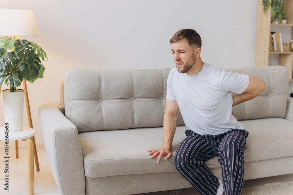Tired bearded man suffering from back pain sitting on couch in living room at home. Male massages painful lower back muscles. Caucasian guy exhausted overworked in apartment.