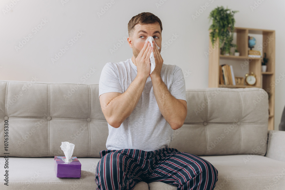 Unhappy middle aged man suffering from fever and flu, blowing nose in napkin, sitting in living room interior. Covid-19 lockdown, treatment of illness, cold and runny