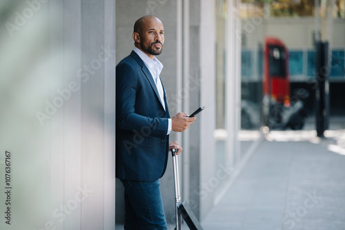 Businessman standing with smart phone near building at footpath photo