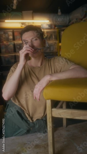 Vertical portrait of proud-looking middle-aged Caucasian woman posing with mustard reupholstered chair in garage workshop, taking off glasses andlooking at camera, small business photo